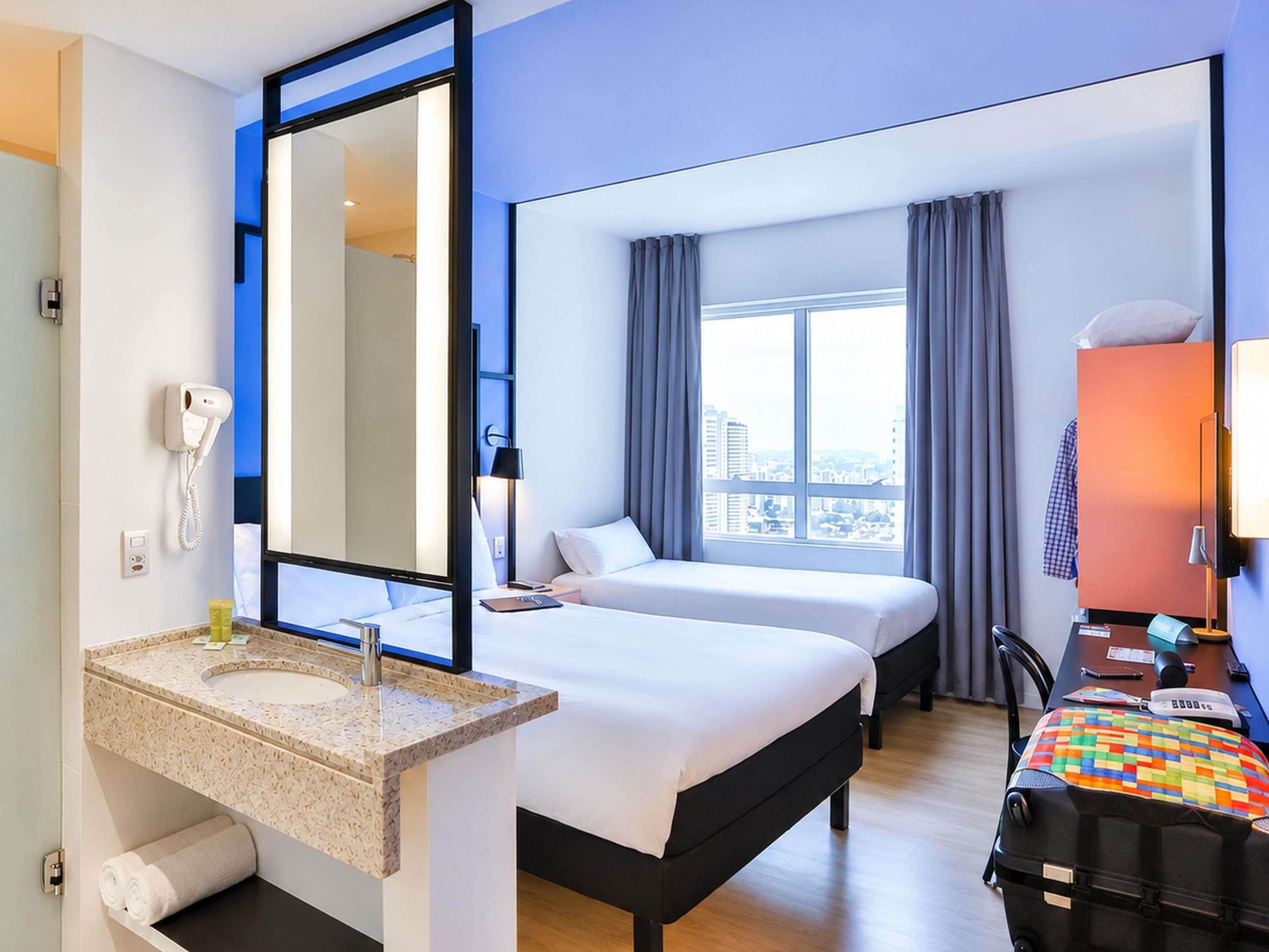 A hotel room with a bed, a desk, a chair, a large window, a mirror, a telephone, and a sink.