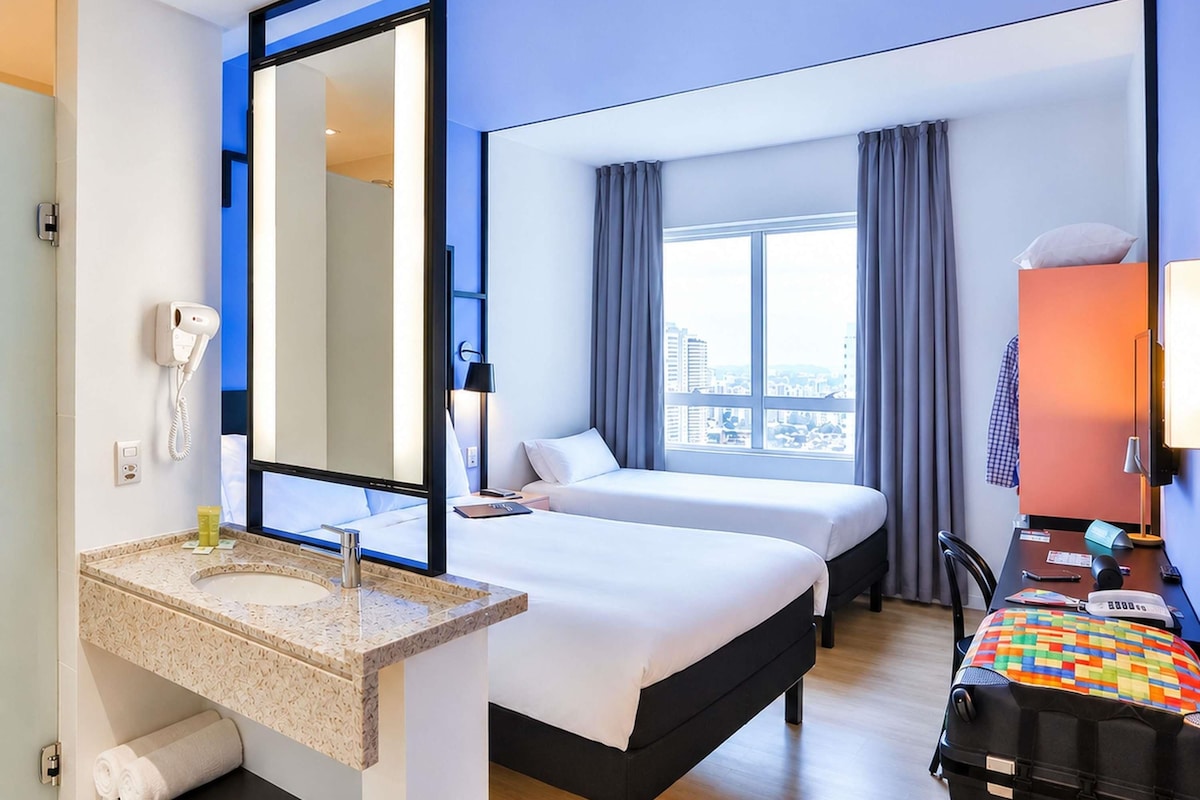 A hotel room with a bed, a desk, a chair, a large window, a mirror, a telephone, and a sink.