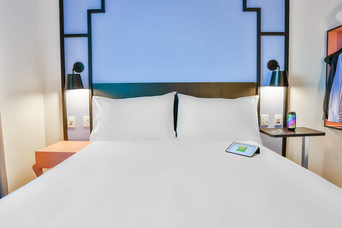Hotelkamer met een bed, nachtkastje, een bedtafel met lamp en wandplank met boek en decoratie.