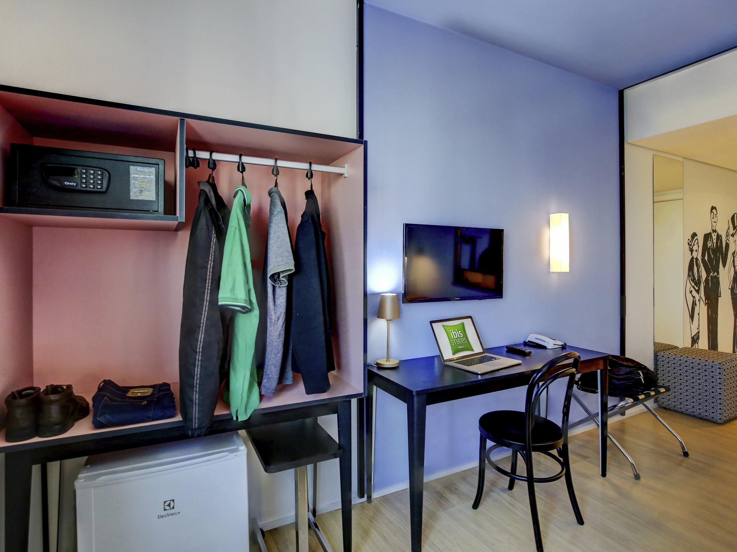 Una habitación con escritorio, silla, televisor y perchero con ropa colgada.