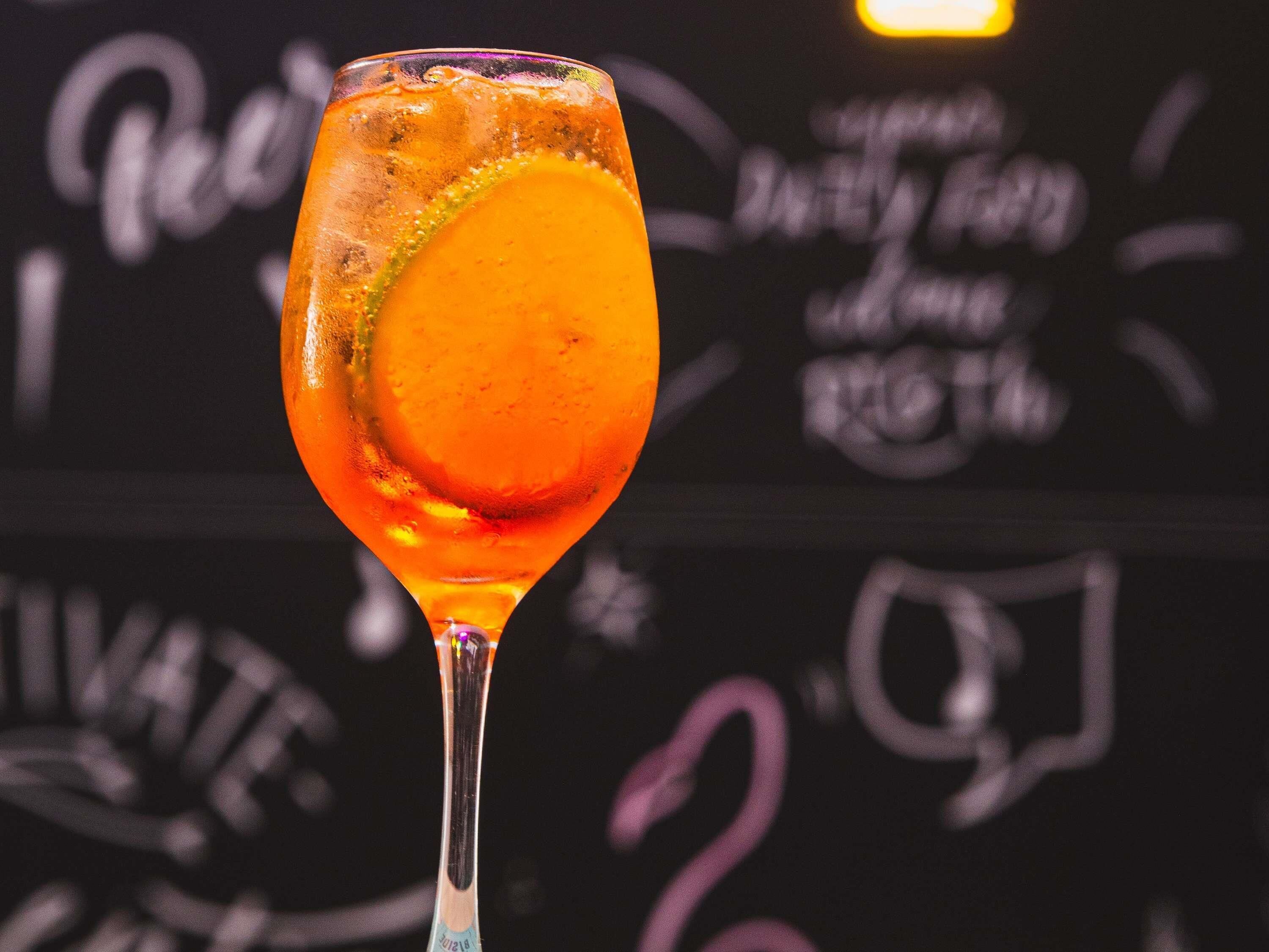 A cocktail glass with a vibrant orange drink, a lime wedge, and a straw, set against a dark background with chalkboard-style writing.