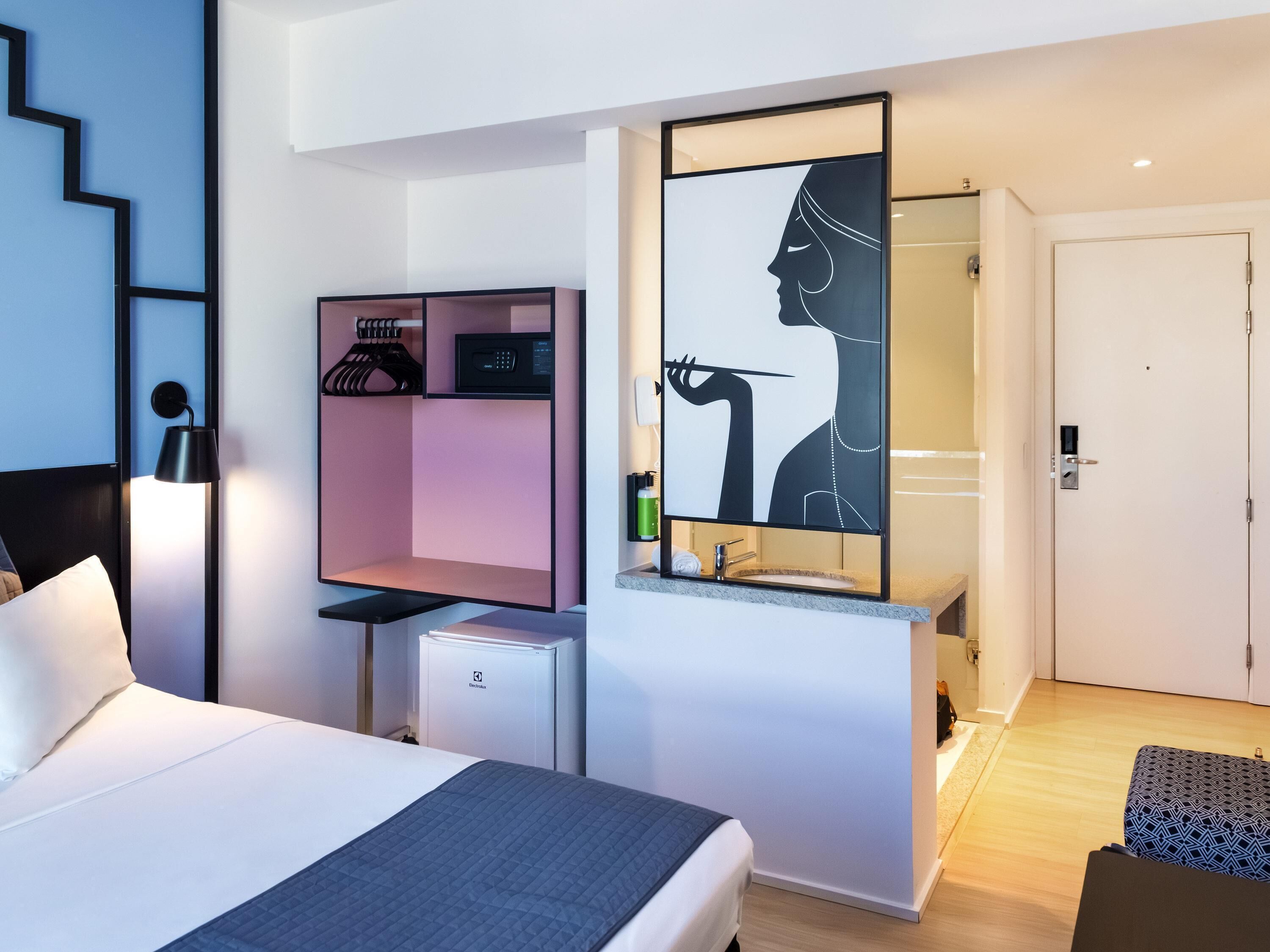 A modern hotel room with a bed, a desk, a mirror, and a wardrobe.