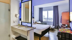 A hotel room with a large bed, a desk, a chair, a desk lamp, a phone, a mirror, and a window with a city view. - ibis Styles Goiania Marista (Goiania)