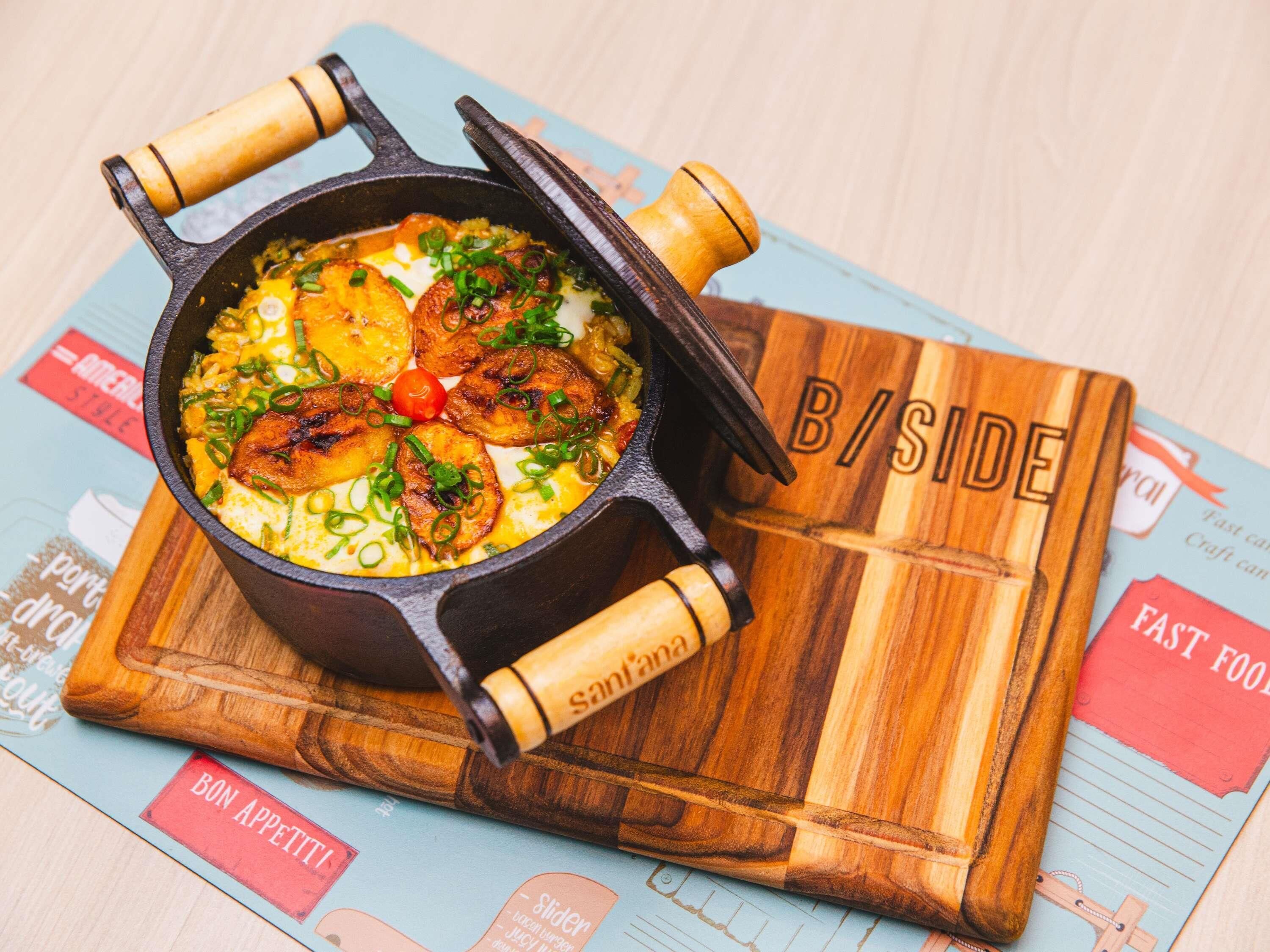 A cast iron skillet with food on a wooden board labeled "B/SIDE".
