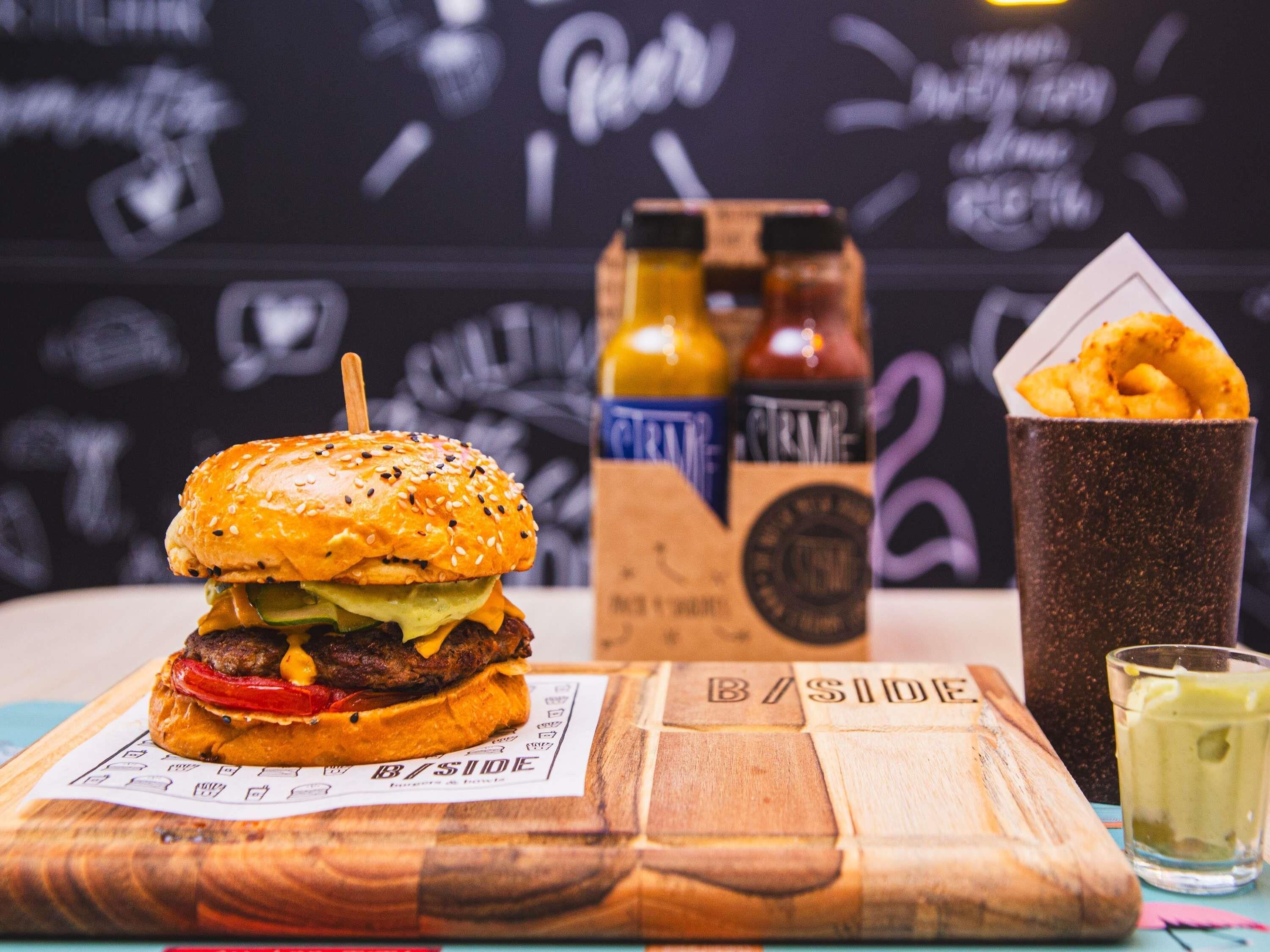 Una hamburguesa sobre una tabla de madera, acompañamientos y una bebida en una mesa.