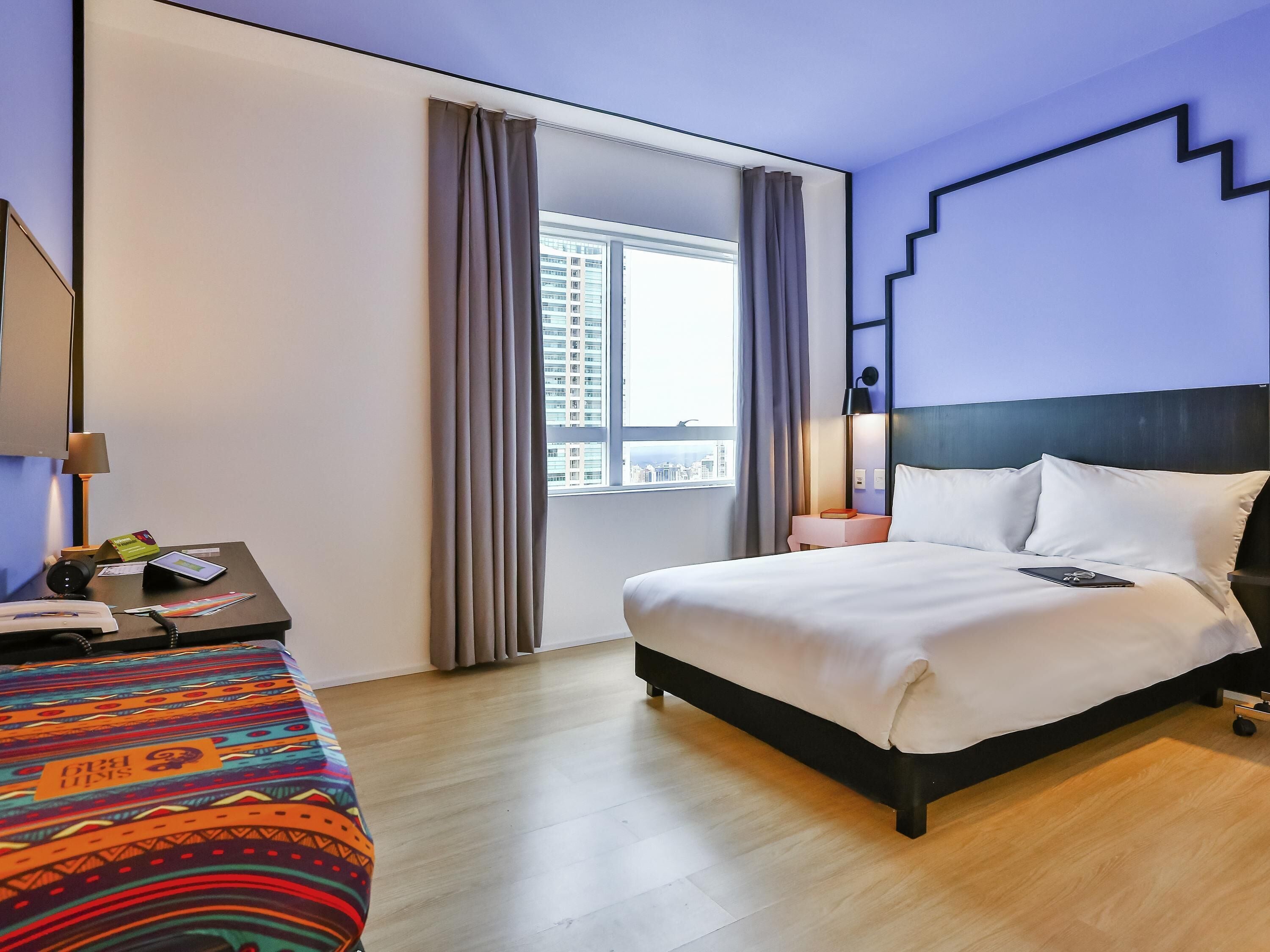 A hotel room with a bed, bedside table, lamp, and a view of the city through the window.