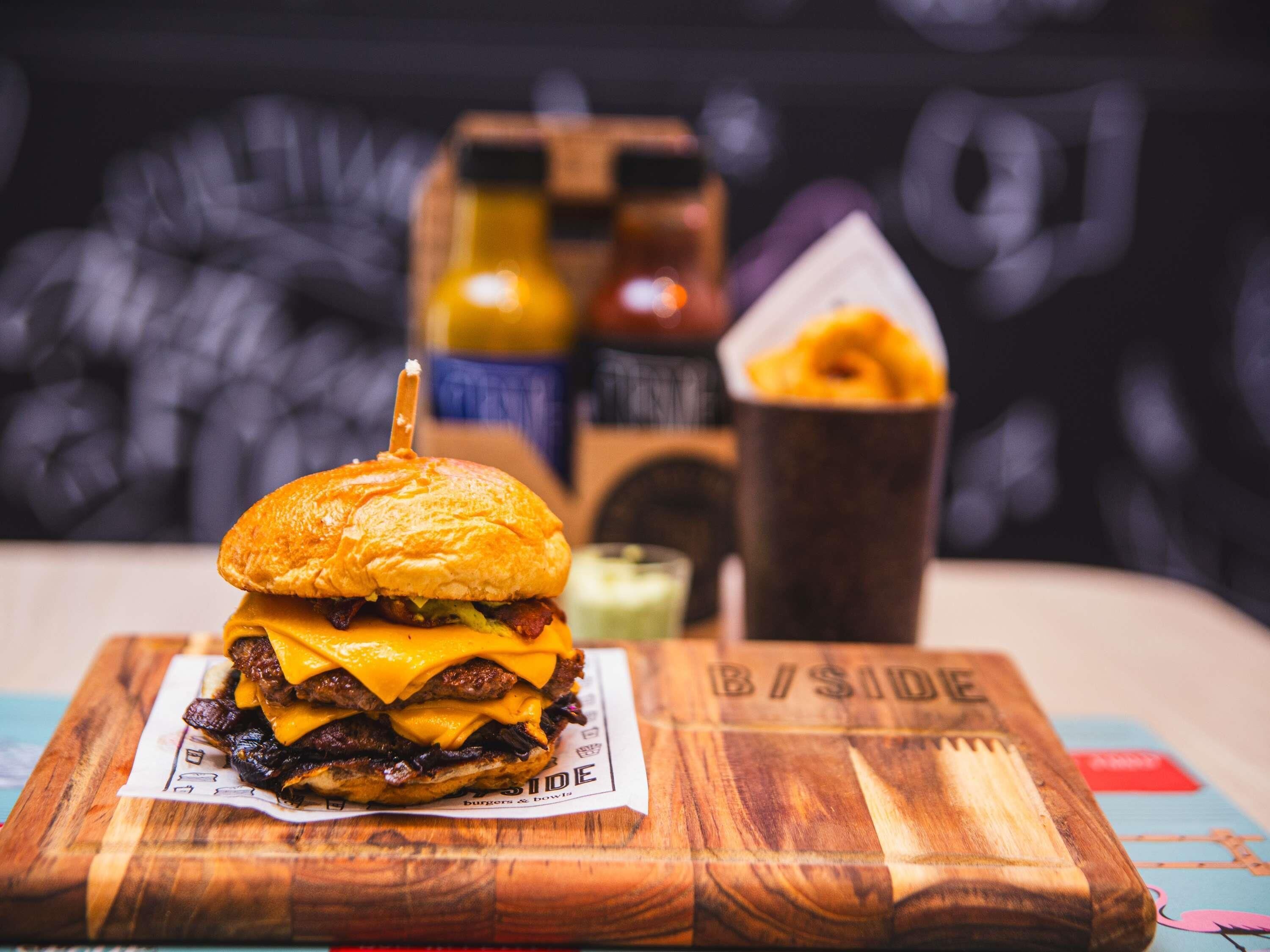 A burger on a wooden board with a napkin, a drink cup, and condiments in the background.