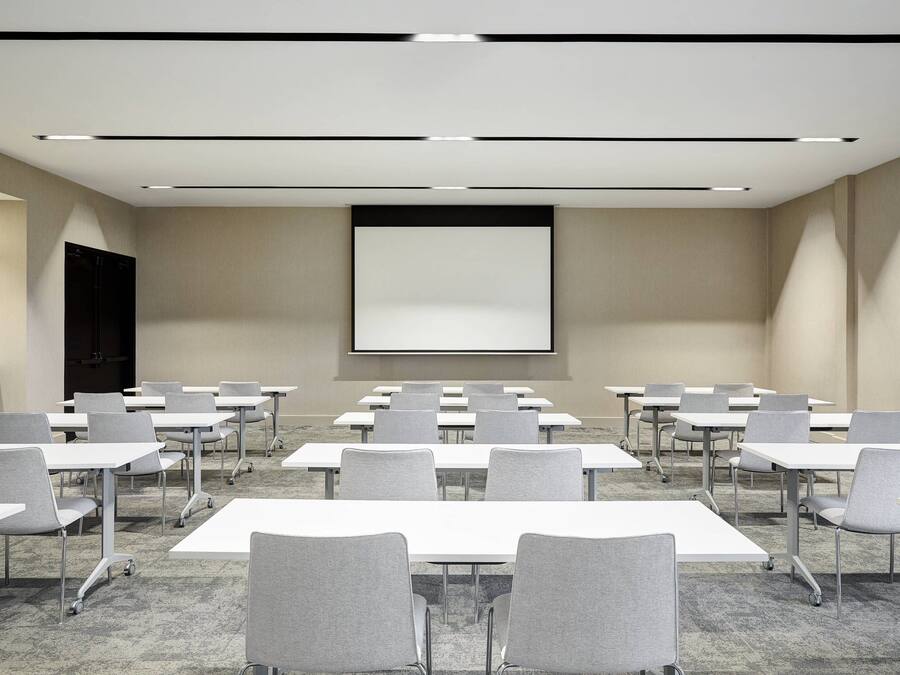 A conference room with rows of tables and chairs, a projector screen, and a carpeted floor.