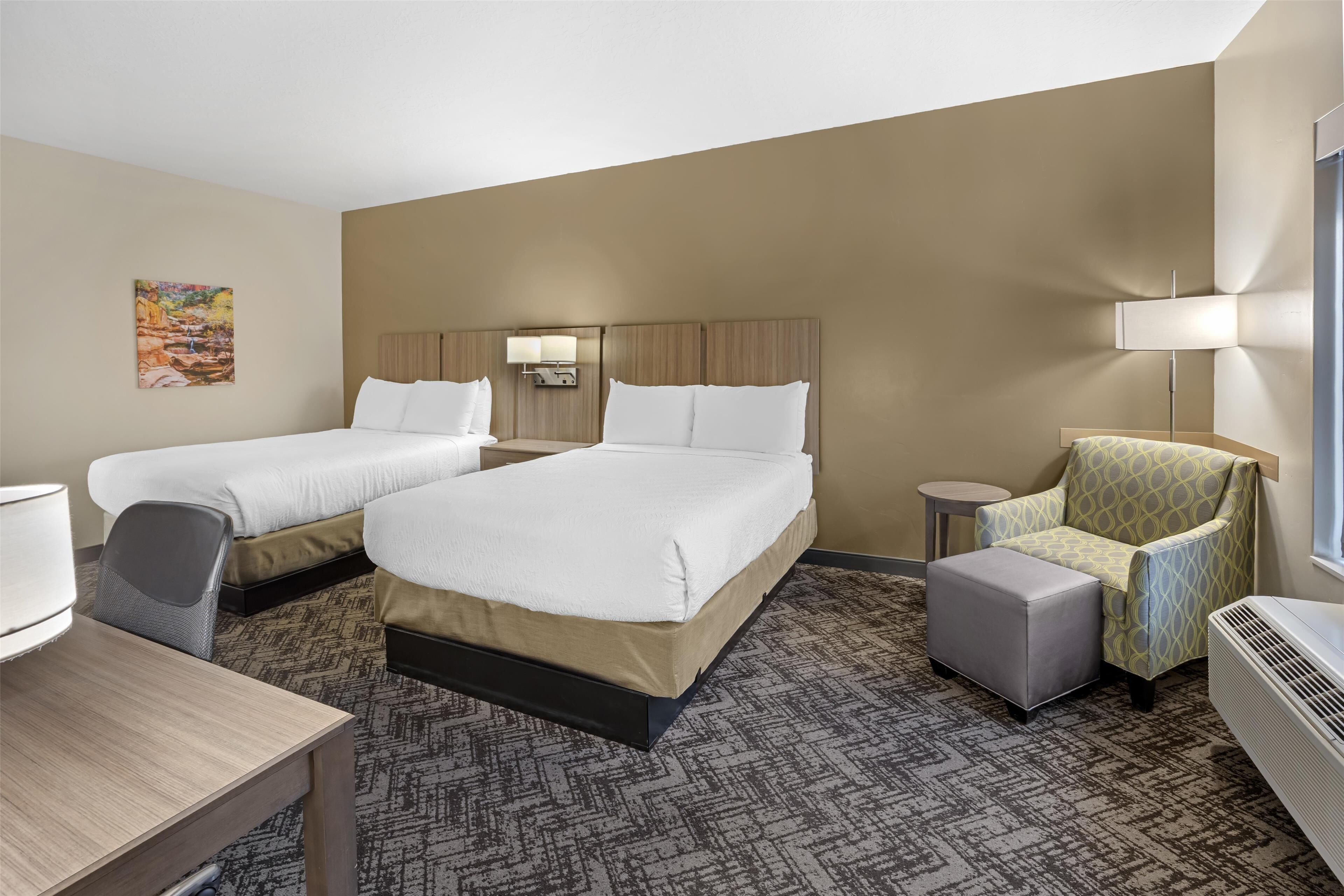 A hotel room with two beds, a desk, a chair, a small table, and a wall-mounted lamp.