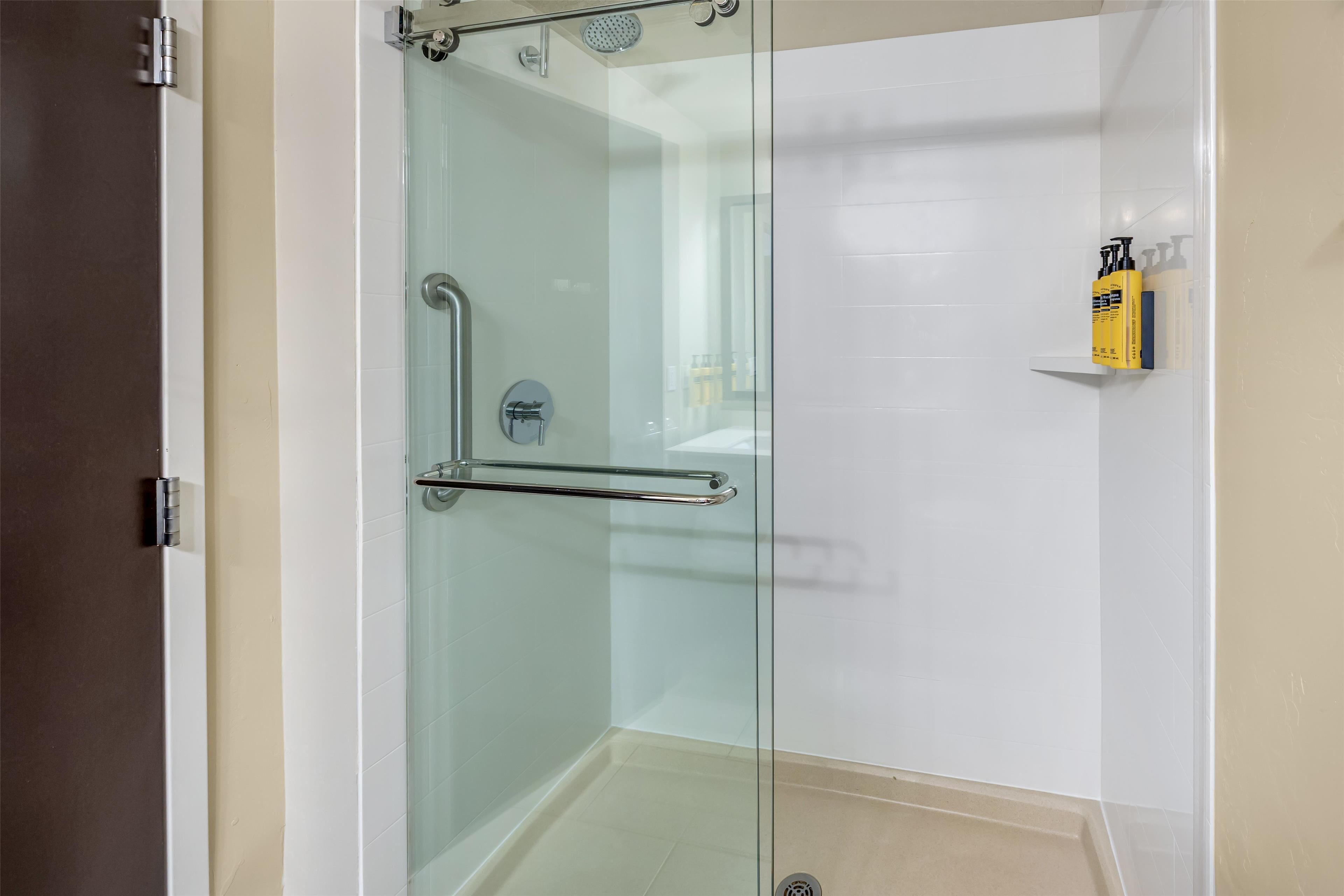 A glass-enclosed shower with a handrail, a white tiled wall, and a shelf with toiletries.