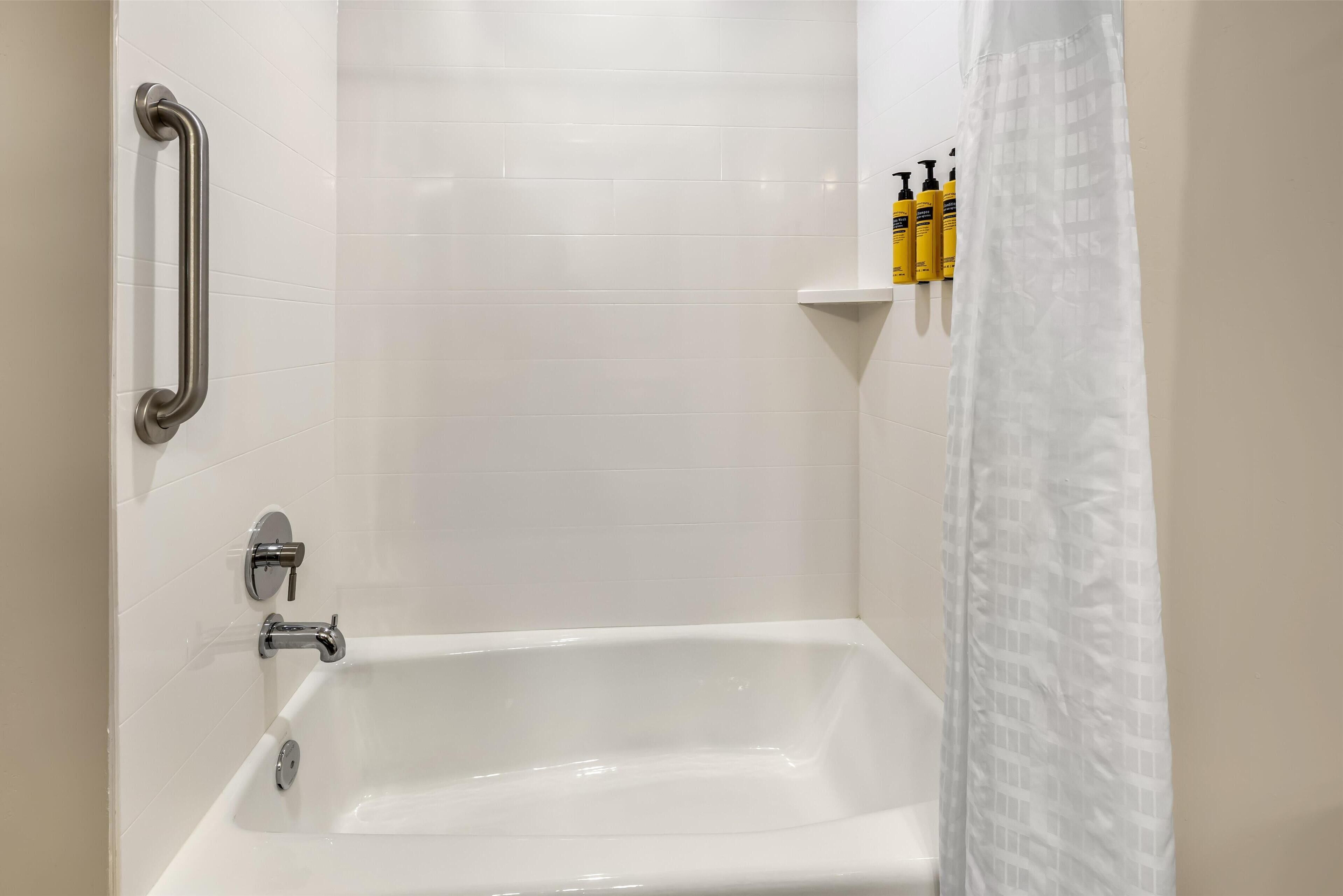 Une salle de bain avec une baignoire blanche, une barre d’appui et une étagère contenant des articles de toilette.