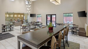 Un grand espace dédié à la salle à manger, avec une longue table, de nombreuses chaises et une plante au centre. On y trouve des luminaires au plafond et un téléviseur fixé au mur.
