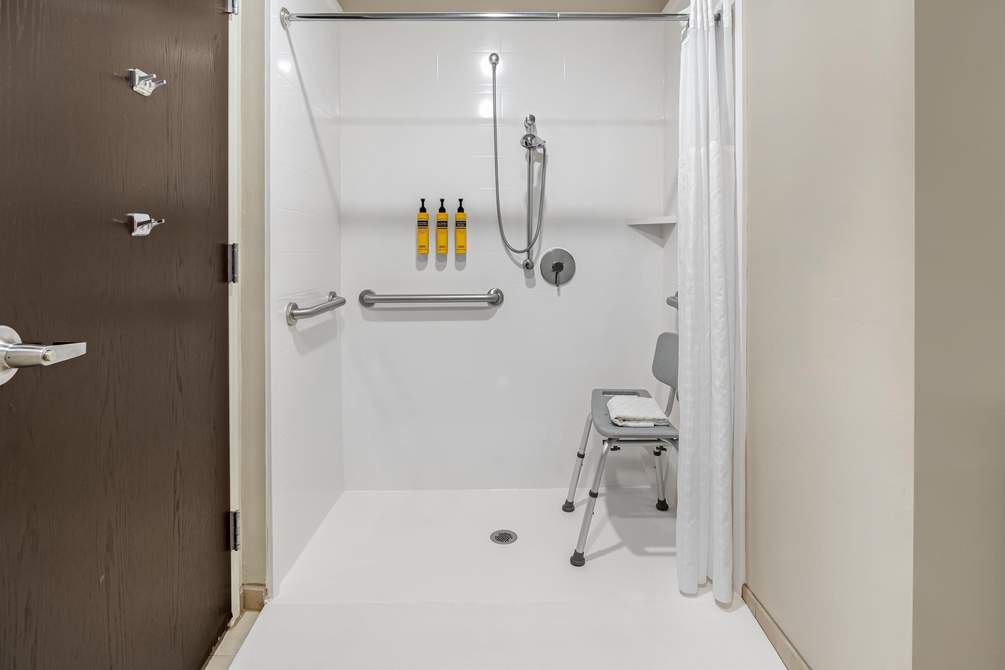 A walk-in shower with a grab bar, a wheelchair-accessible bench, and a hand sanitizer dispenser.