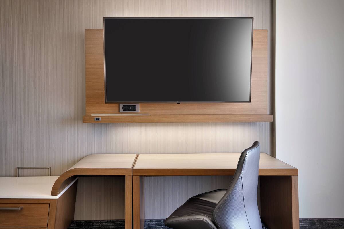 Une chambre d’hôtel moderne avec un bureau en bois, un fauteuil en cuir et un téléviseur à écran plat fixé au mur.