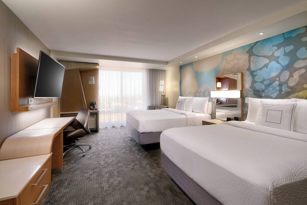 Une chambre d’hôtel avec deux lits, un bureau, un téléviseur à écran plat, une grande fenêtre avec des rideaux légers et une fresque murale abstraite aux couleurs vives.