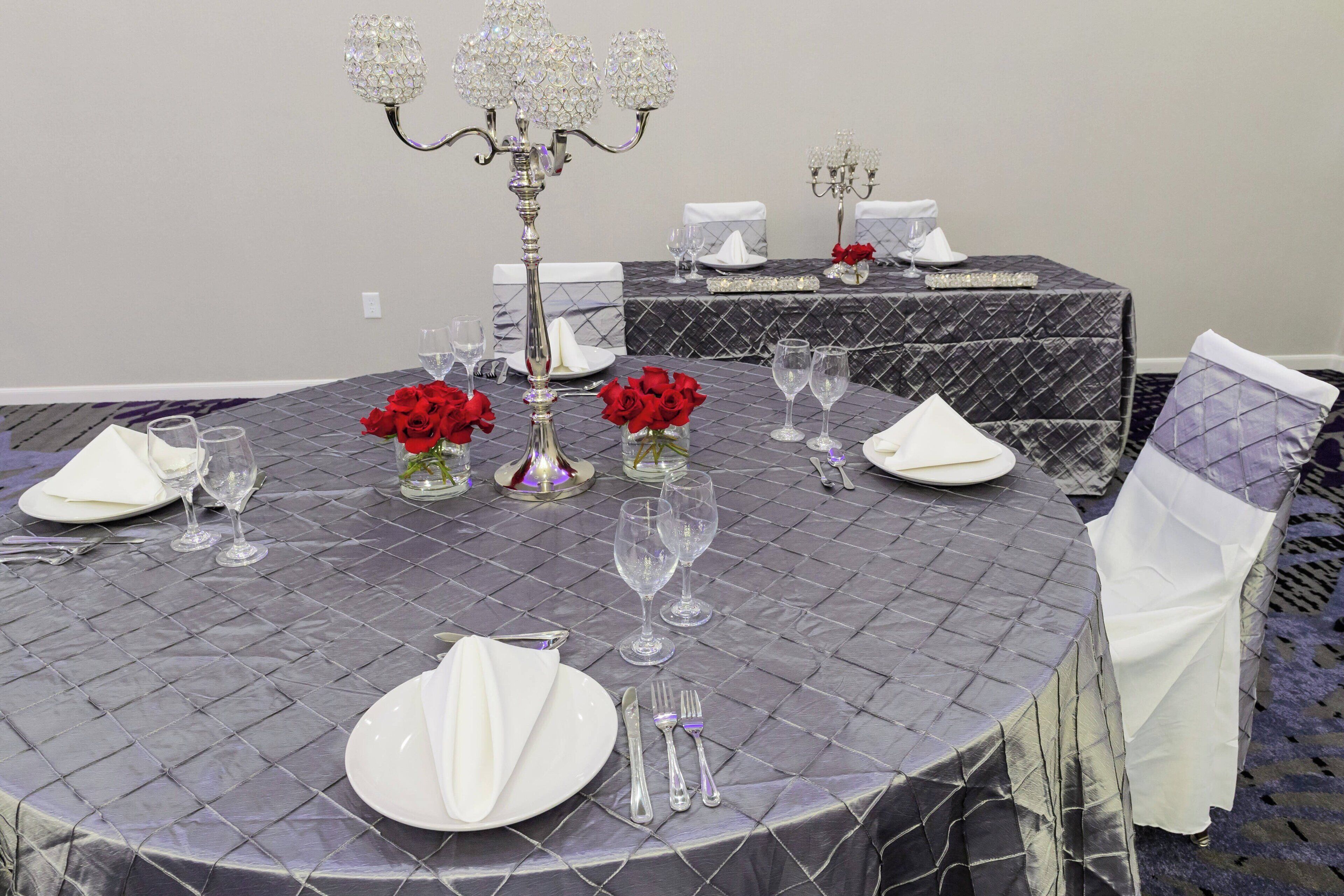 A round dining table with a diamond-patterned tablecloth, white napkins, silverware, and red roses in vases.