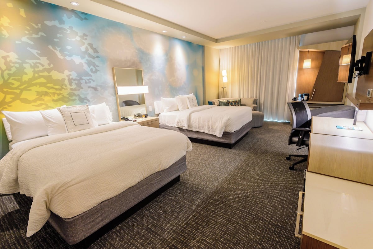 Ein Hotelzimmer mit zwei Betten, einem Schreibtisch, einem Sessel und einem großen Spiegel, sowie einer Wand mit einem farbenfrohen abstrakten Muster.