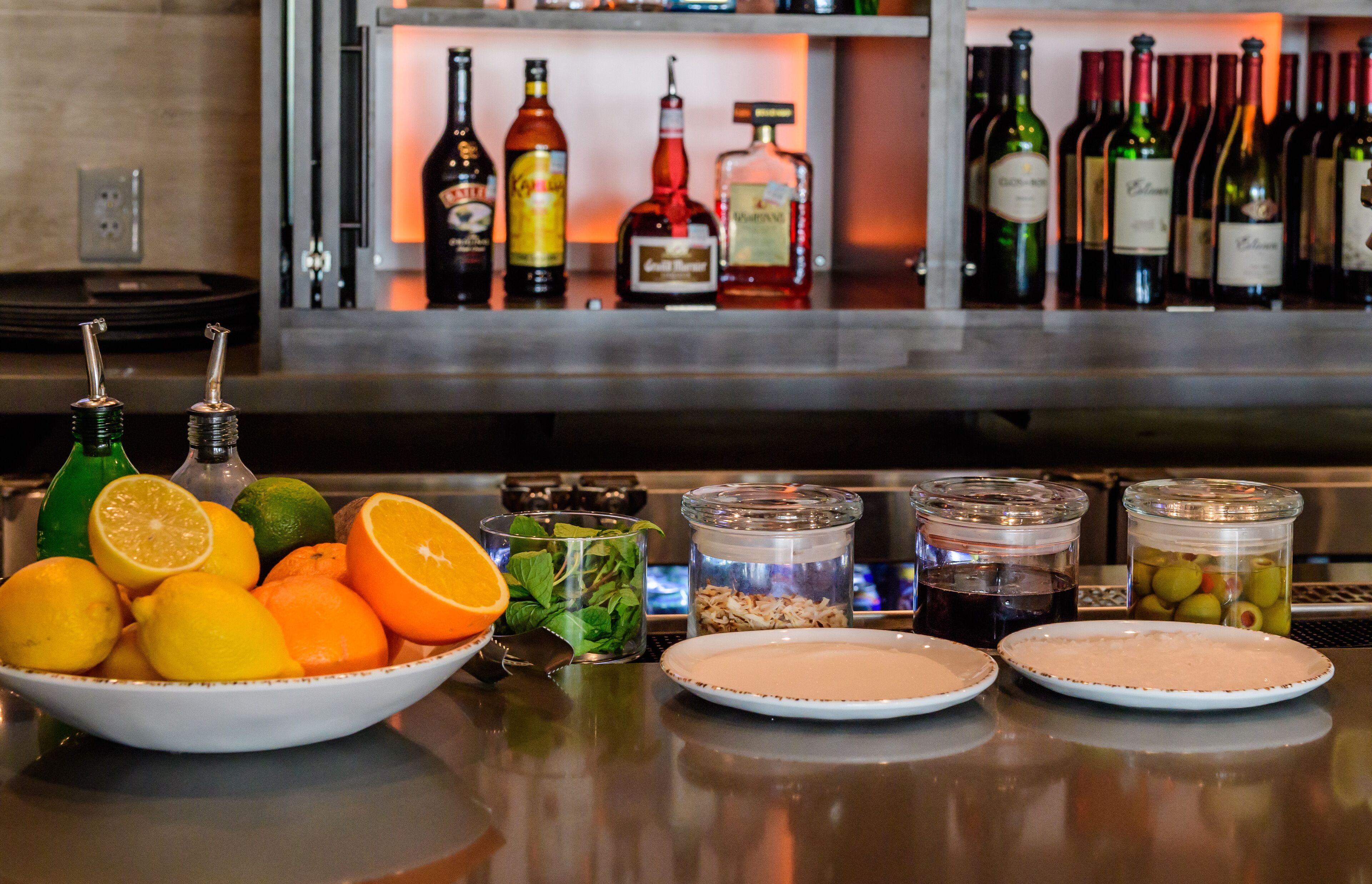 A bar area with bottles of alcohol, a bowl of citrus fruits, and jars of pickles and olives.