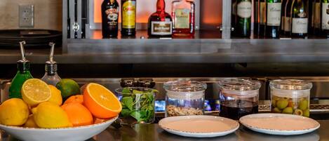 A bar area with bottles of alcohol, a bowl of citrus fruits, and jars of pickles and olives.
