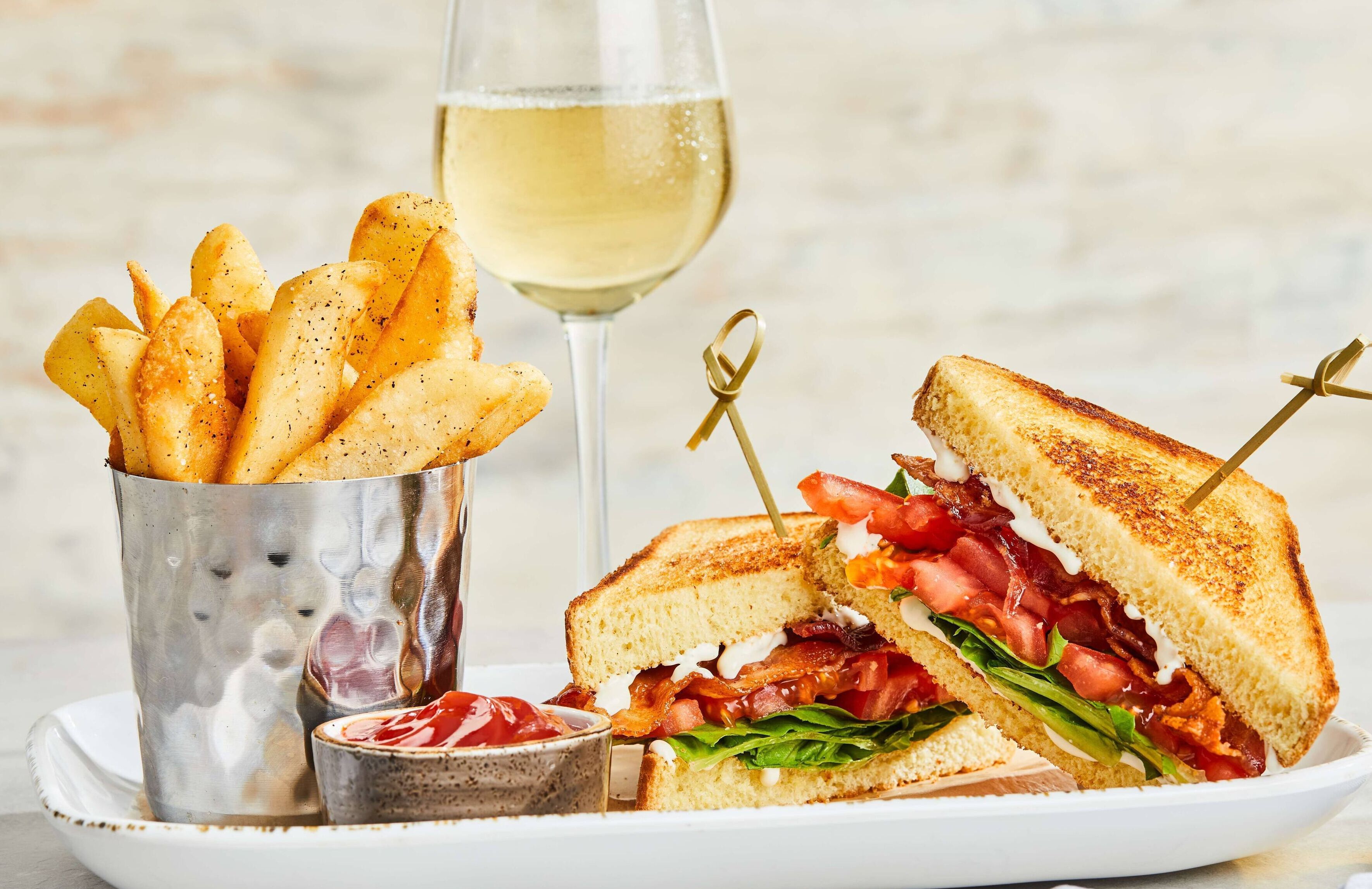 A sandwich with bacon, tomato, and lettuce, a glass of white wine, and a cup of fries.