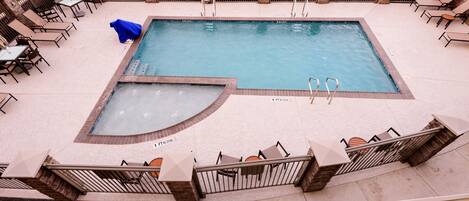 A rectangular pool with a diving area, surrounded by lounge chairs and tables, and a stone wall with a warning sign.