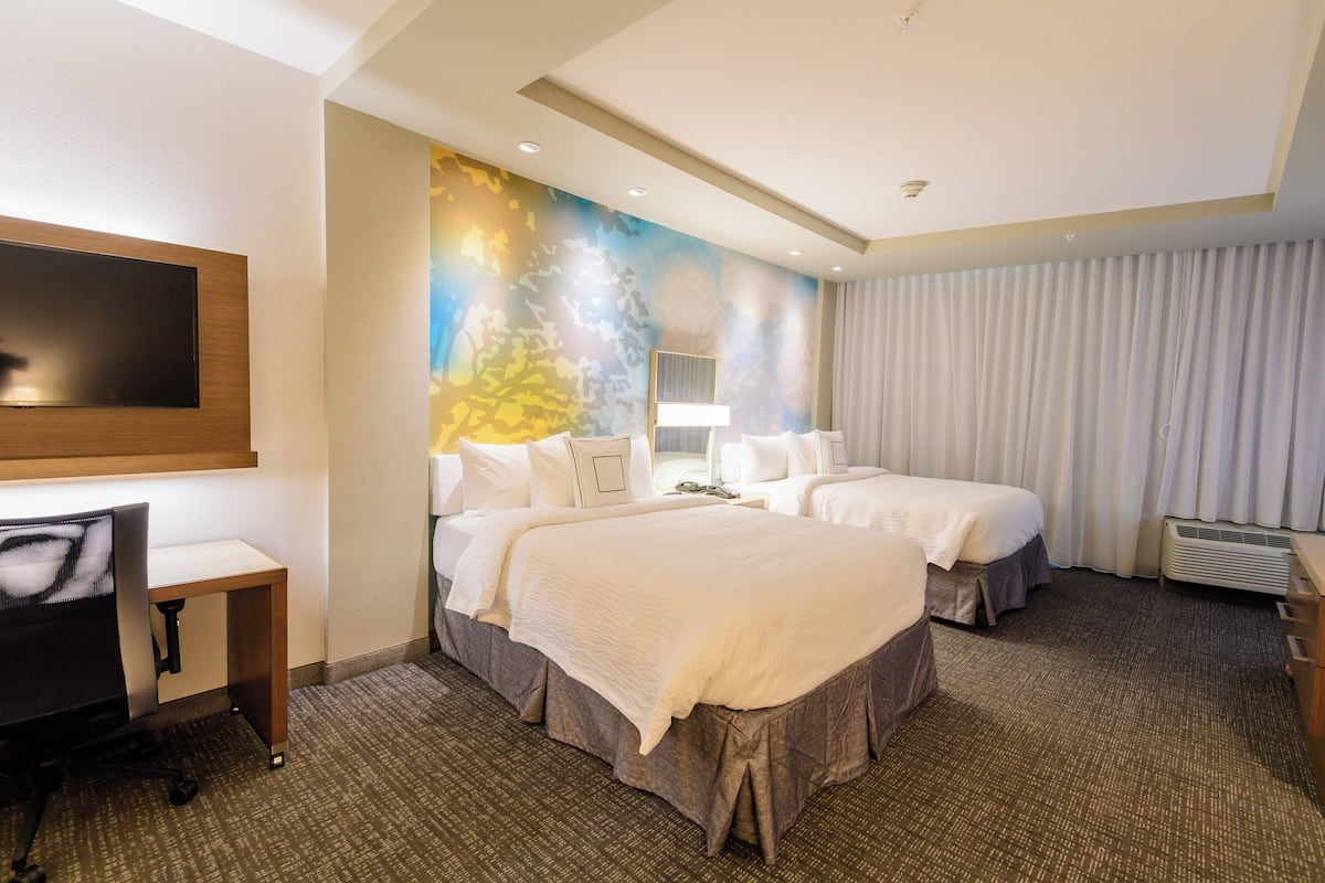 Ein Hotelzimmer mit zwei Betten, einem Schreibtisch, einem Fernseher und einem großen Fenster mit Vorhängen.