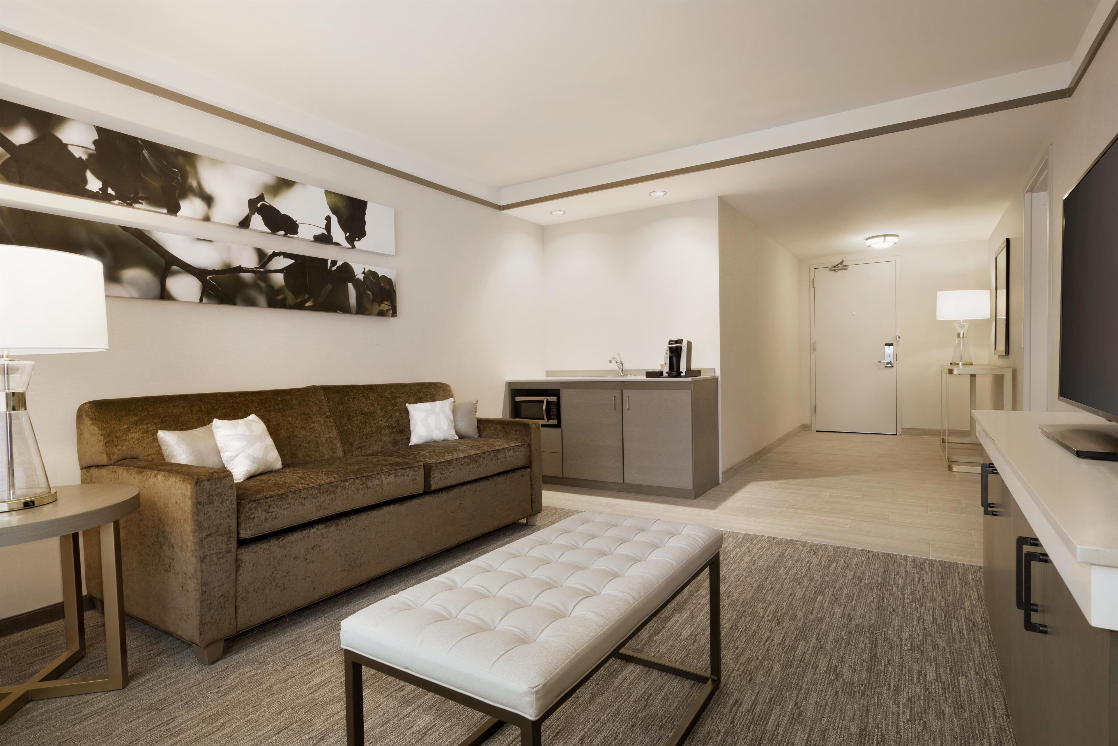 A modern hotel room with a sofa, a coffee machine, a TV, and a small kitchenette.