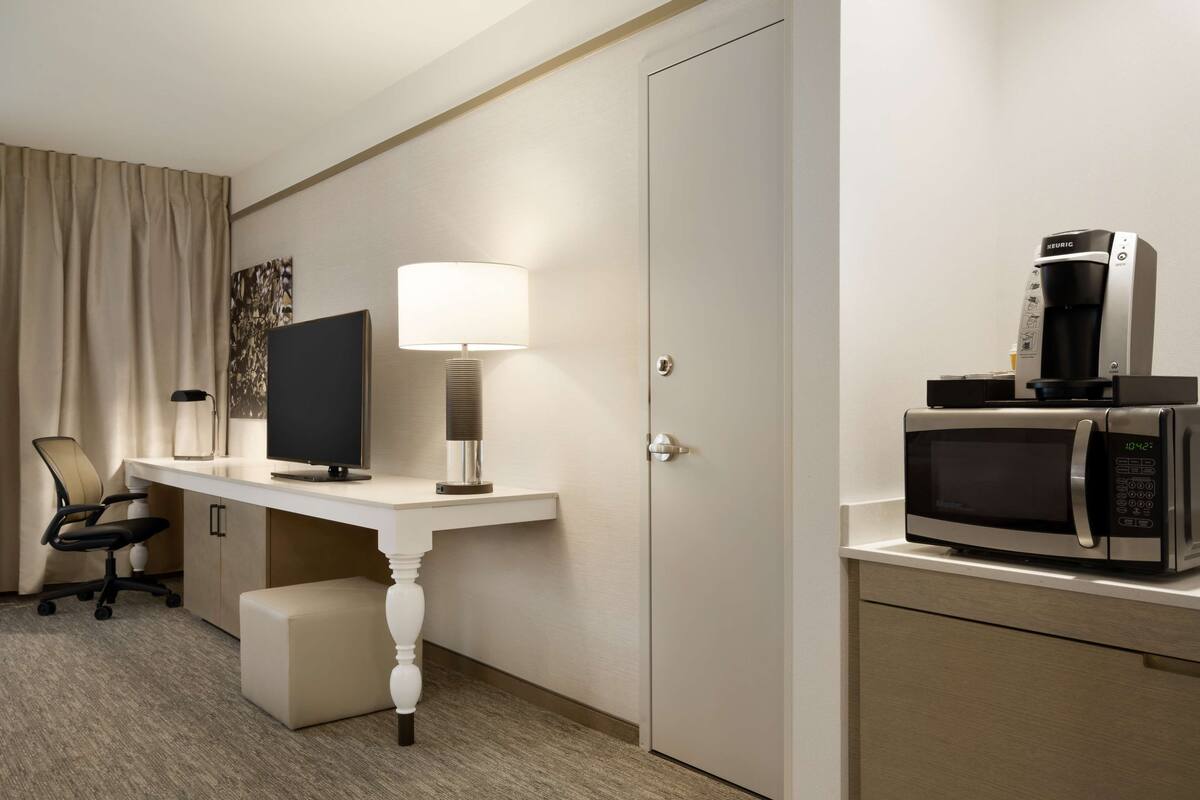 A hotel room with a desk, a computer, a lamp, a chair, a coffee maker, and a microwave.