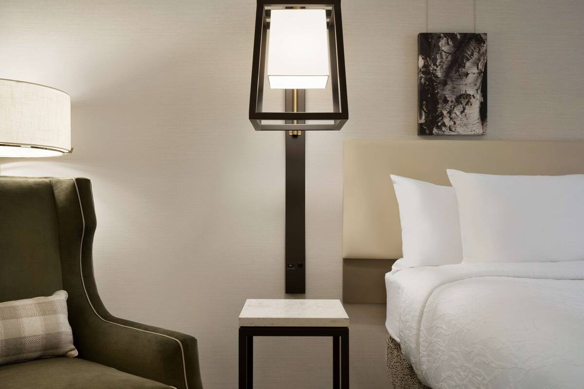 A hotel room with a bed, a nightstand, a lamp, a chair, and a painting on the wall.