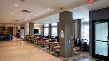 A hotel lobby with a long corridor, multiple dining tables, and chairs, a large pillar, and an exit sign.
