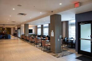 A hotel lobby with a long corridor, multiple dining tables, and chairs, a large pillar, and an exit sign. - Hilton Garden Inn Newtown Square Radnor (Newtown Square)