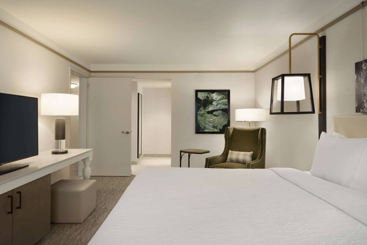 A hotel room with a large bed, a desk with a TV, a chair, a small table, and a painting on the wall.