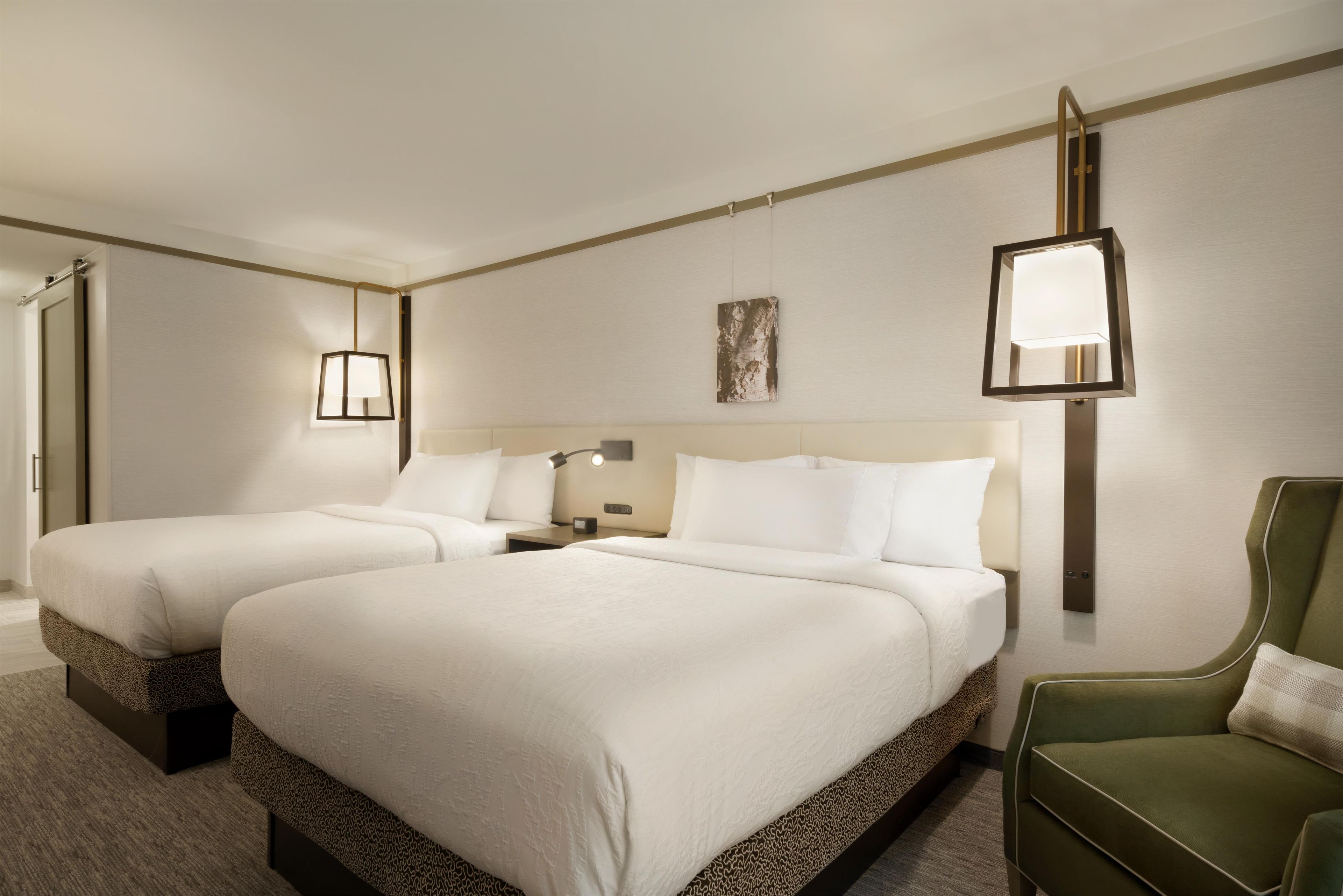A hotel room with two beds, a nightstand, a lamp, a painting, and a green armchair.