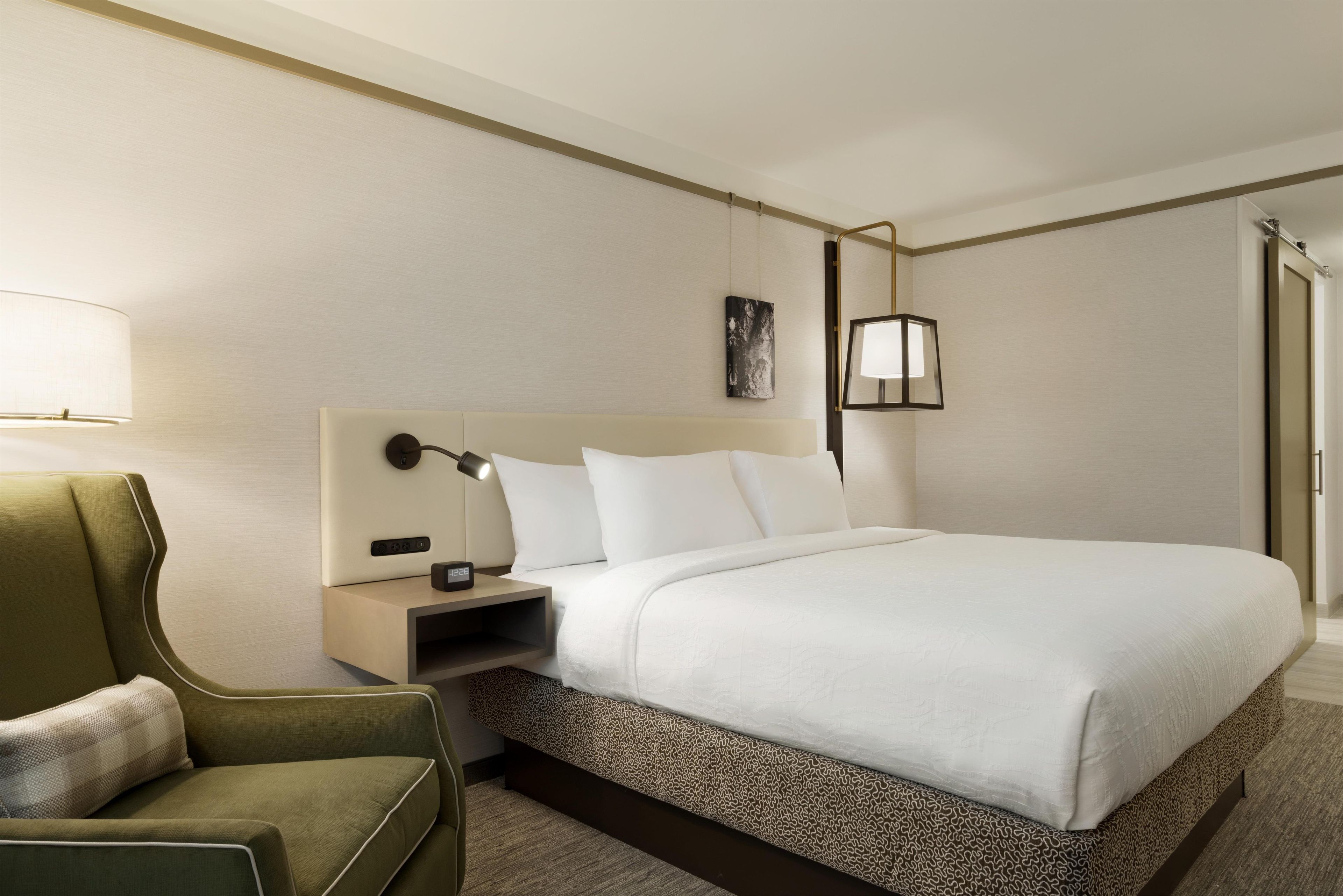 A hotel room with a bed, a nightstand, a lamp, a chair, and a painting on the wall.