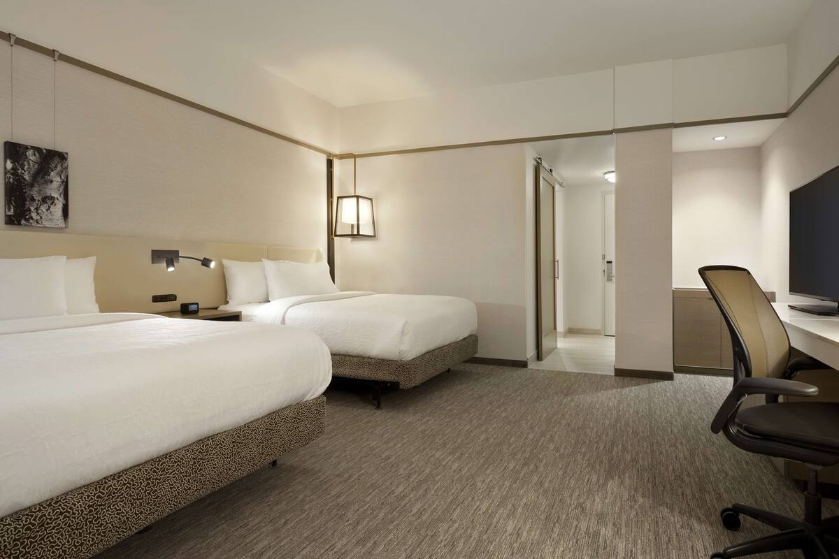 A hotel room with two beds, a desk, a chair, and a lamp.