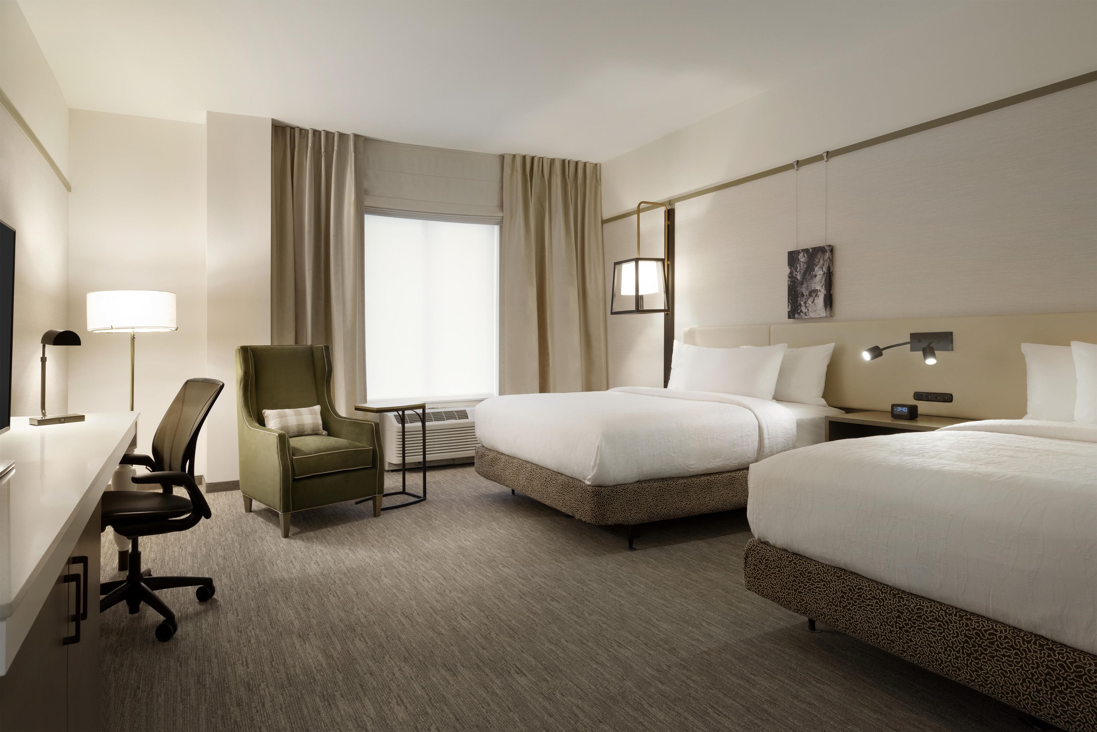 A hotel room with two beds, a desk, a chair, a lamp, and a painting on the wall.