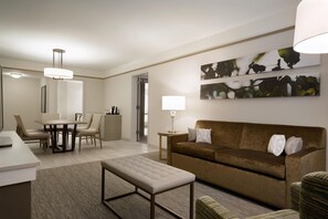 A modern living room with a brown sofa, a white ottoman, a dining table with chairs, and a kitchen area in the background. - Hilton Garden Inn Newtown Square Radnor (Newtown Square)