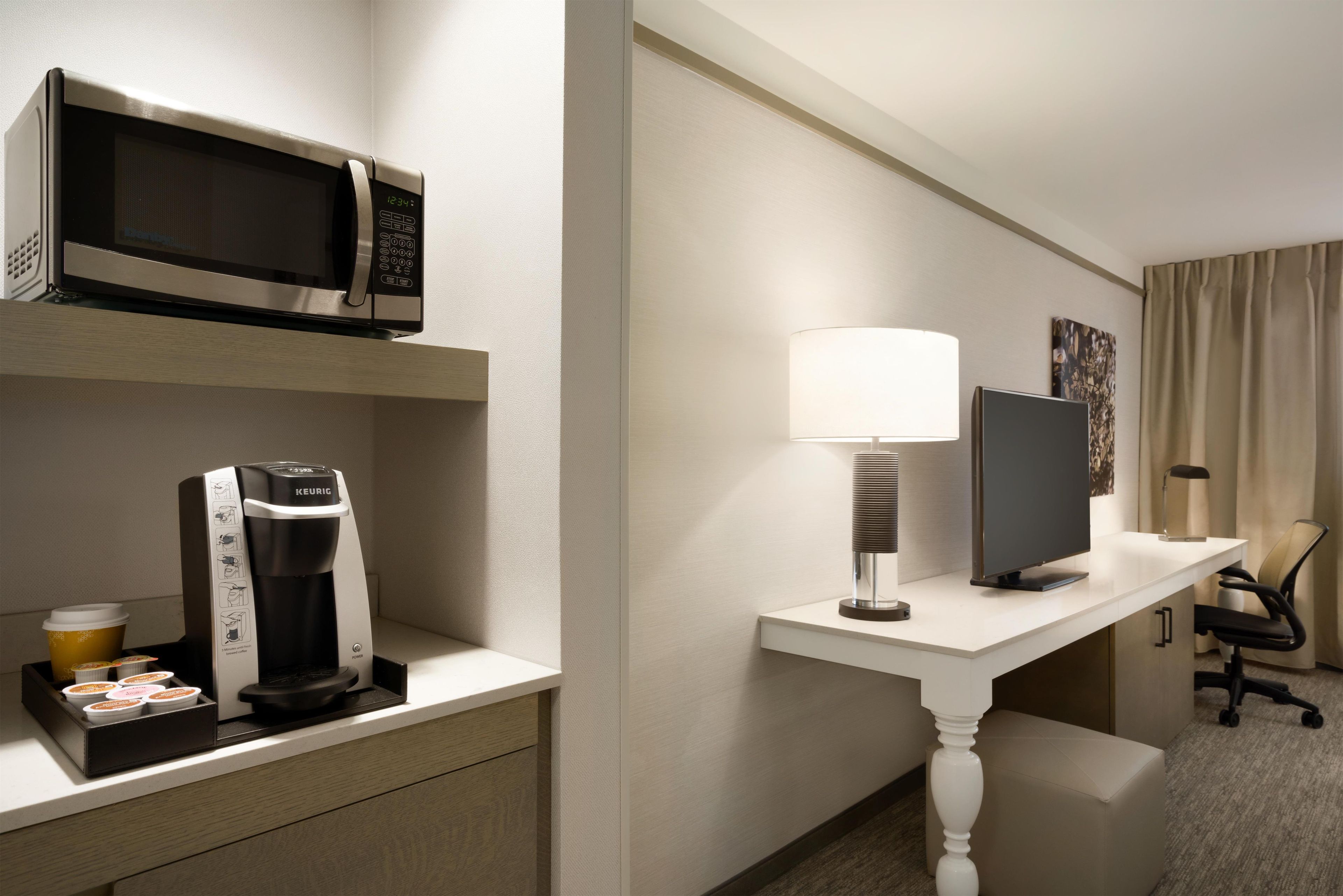 A hotel room with a microwave, coffee maker, desk with a lamp, TV, and a chair.