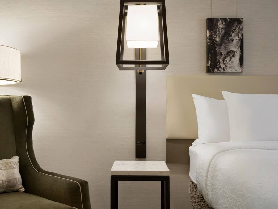 A hotel room with a bed, a nightstand, a lamp, a chair, and a painting on the wall.