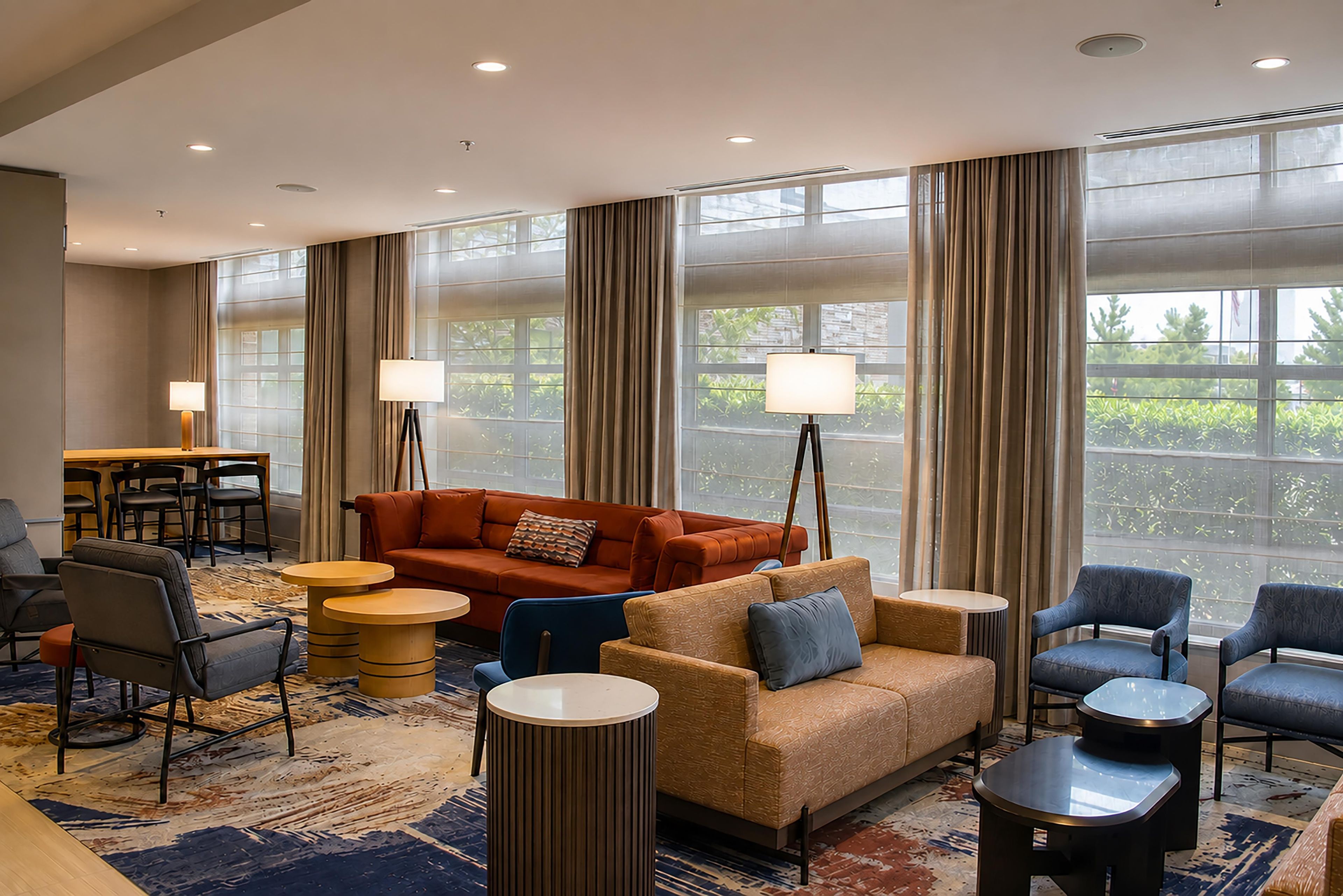 A hotel lobby with a seating area, including a sofa, chairs, and a coffee table. There are large windows with curtains, a dining area, and a lamp.