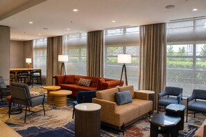 Un hall de hotel con un área de descanso, que incluye un sofá, sillas y una mesa de centro. Hay ventanas amplias con cortinas, un comedor y una lámpara.