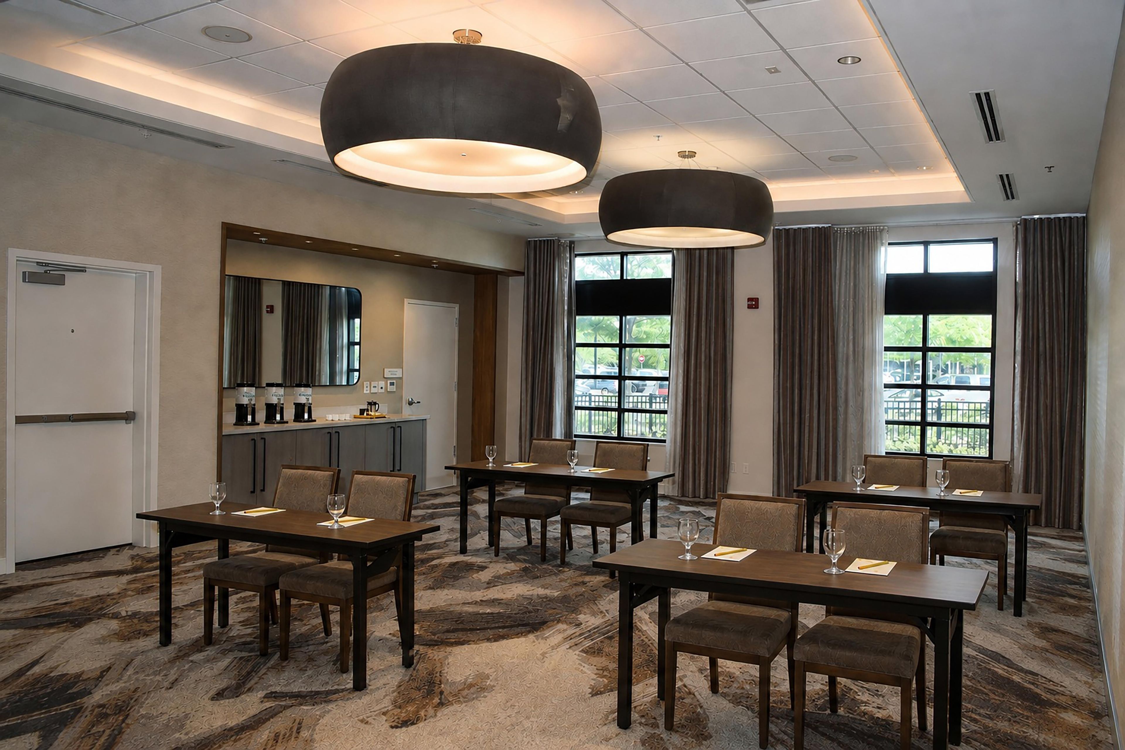 Habitación de hotel con zona de comedor, minibar y ventanas grandes con cortinas.