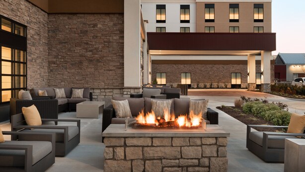An outdoor seating area with a stone fire pit and modern furniture, surrounded by a building with a stone facade and multiple windows. - Hilton Garden Inn Newtown Square Radnor (Newtown Square)