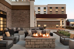 Un espacio al aire libre con una chimenea de piedra y mobiliario moderno, rodeado por un edificio de fachada de piedra y múltiples ventanas.