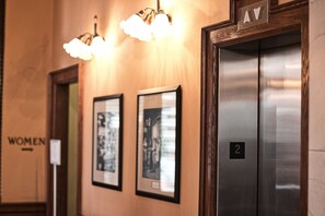 Ein Aufzug mit der Beschriftung '2', eine Wand mit gerahmten Bildern und ein Schild mit der Aufschrift 'FRAUEN'.