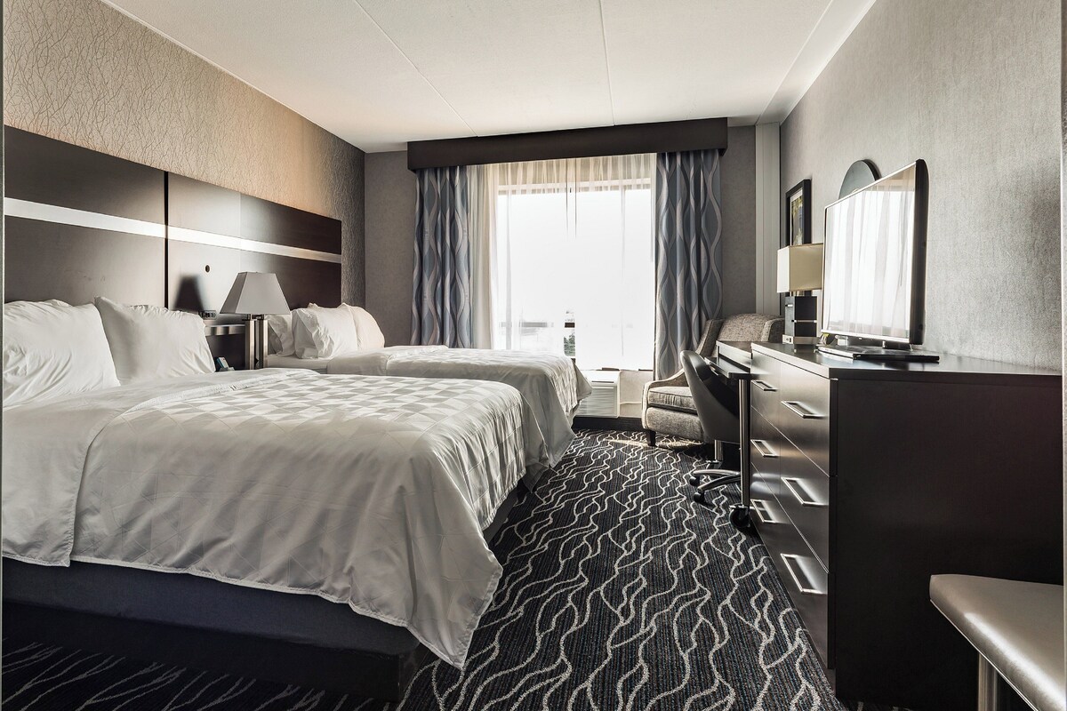 Ein Hotelzimmer mit zwei Betten, einem Schreibtisch, einem Sessel, einem Fernseher und einem großen Fenster mit Vorhängen.