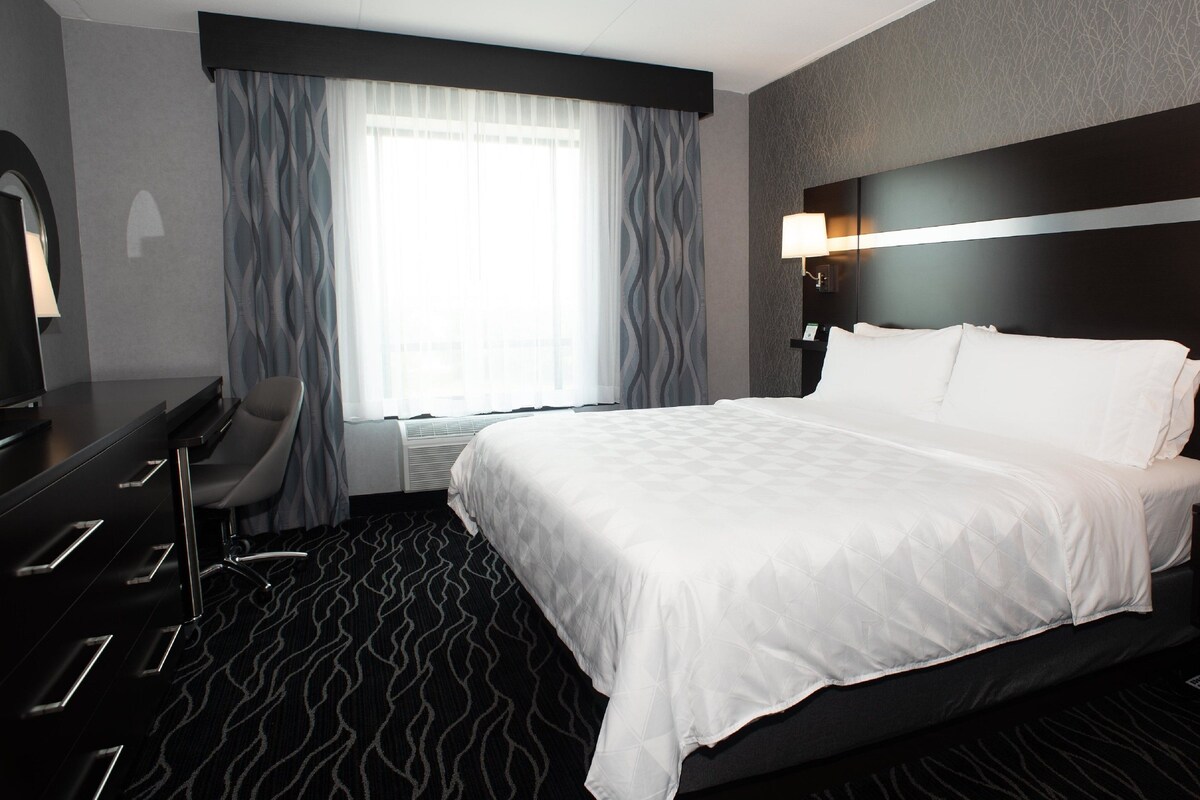 Ein Hotelzimmer mit einem großen Bett, einem Schreibtisch mit Stuhl, einer Schreibtischlampe und einem Fenster mit Vorhängen.