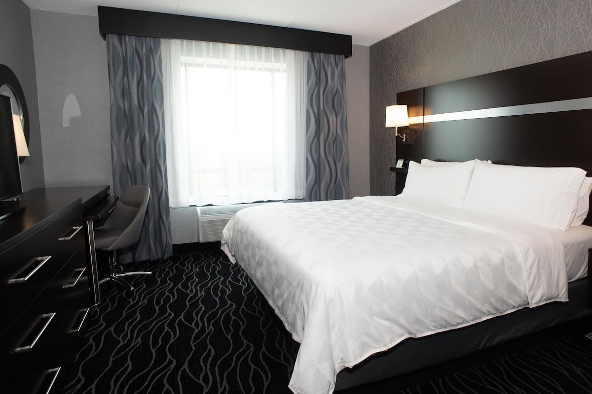 A hotel room with a large bed, a desk with a chair, a desk lamp, and a window with curtains.