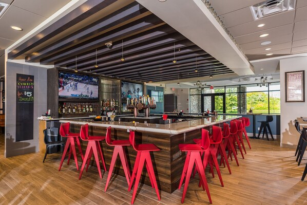 Zona bar moderna con un lungo bancone, sgabelli rossi e un grande schermo che mostra una partita sportiva.