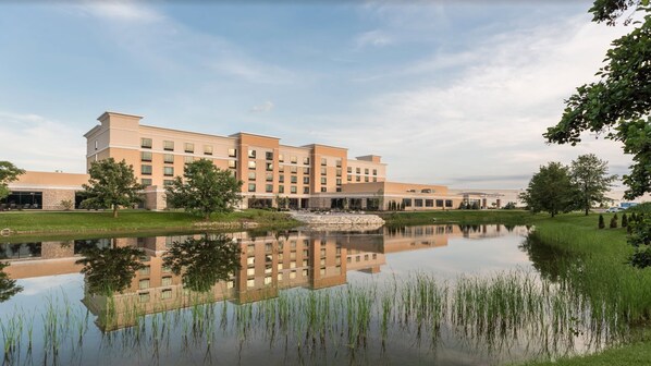 A multi-story hotel with a reflection in a calm pond, surrounded by greenery and a clear sky.